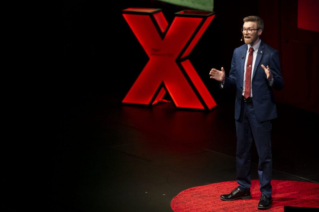 Malachi Rein on Ted Talk stage with a red illuminated X in the background